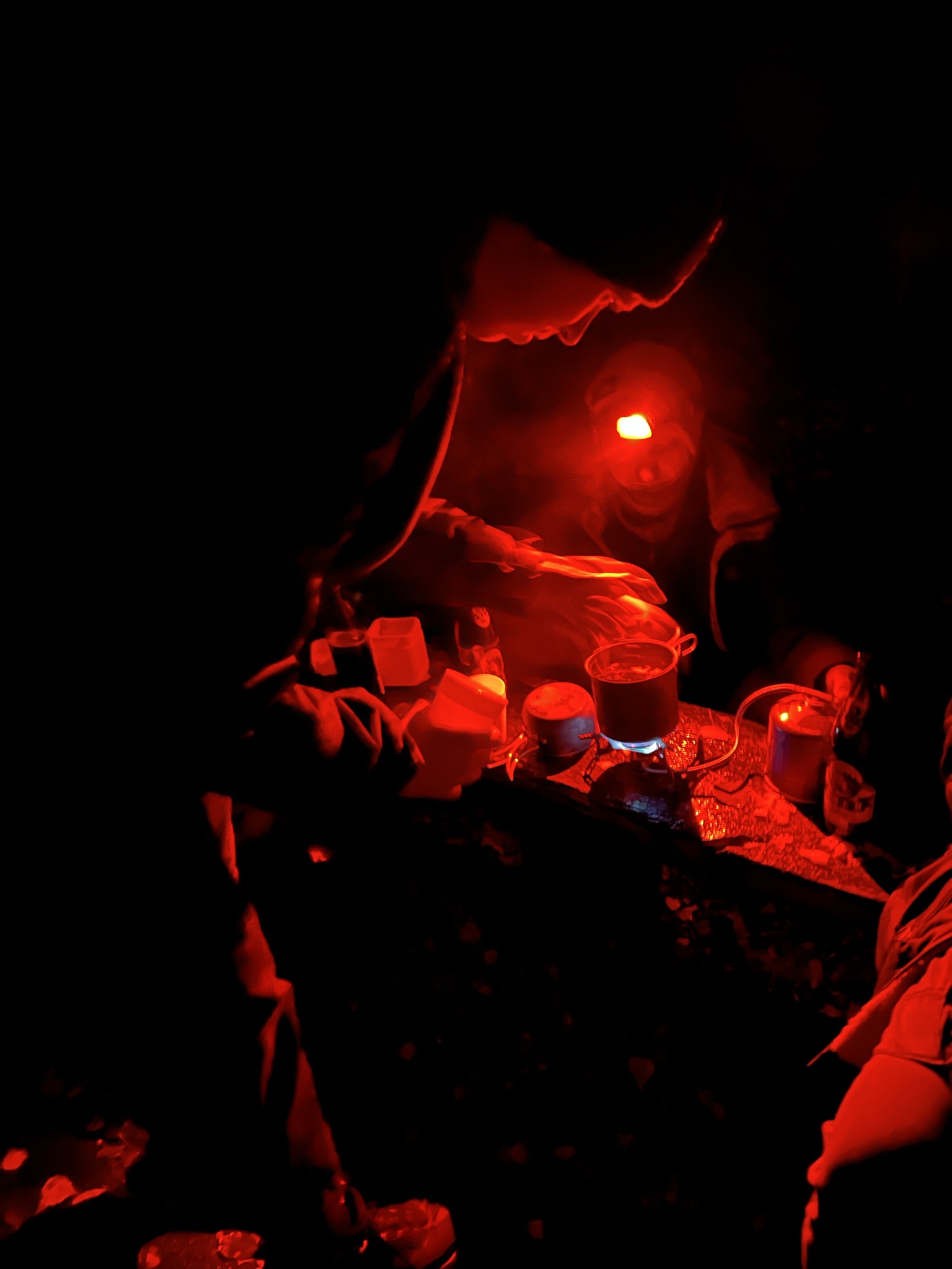People making hot drinks with a gas cooker at night, lit by red headlamps.